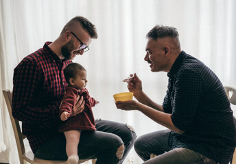 Male gay couple with adopted baby girl at home - Two handsome dads feed the baby girl on kitchen -...