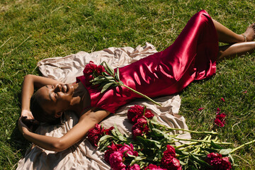 Portrait of a black girl lying in a grass in a silk dress with a peony flowers