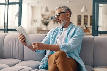 Busy senior man in casual clothing using digital tablet while sitting on the sofa at home