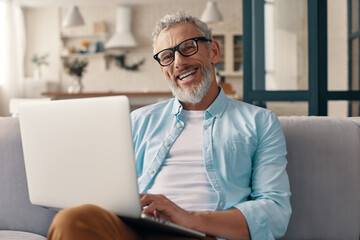 Senior man in casual clothing using laptop and looking at camera with smile while sitting on the sofa at home - obrazy, fototapety, plakaty