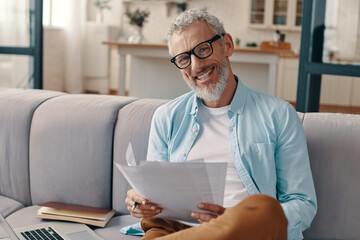 Happy senior man checking the papers and looking at camera with smile while sitting on the sofa at...