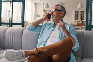 Modern senior man in casual clothing and eyeglasses talking on the smart phone while sitting on the sofa at home