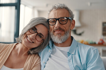 Senior couple in casual clothing smiling and looking at camera while spending time at home