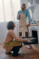 Cheerful senior couple in aprons preparing healthy dinner in oven and smiling while spending time...