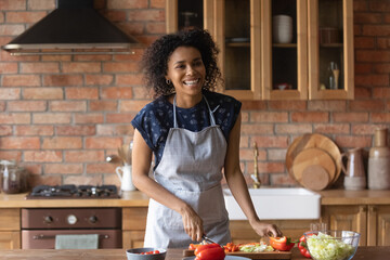 Happy housewife. Laughing millennial black woman in apron enjoy cooking favorite healthy food dishes at modern home kitchen. Joyful young afro american lady cut fresh vegetable salad for family lunch - Powered by Adobe