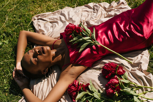 Portrait of a beautiful black girl lying on the grass in a silk dress with a peony flowers