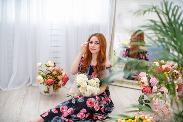 Attractive young woman in floral print dress sits among bouquets flowers in an elegant floral interior. pretty girl with curly red hair, in beautiful dress, in floral interior, smiles. spring festival