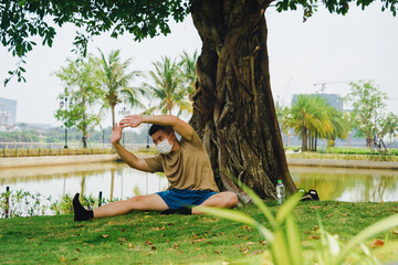 Asian man practicing meditation with wear protective mask on face, yoga exercise under big tree in...