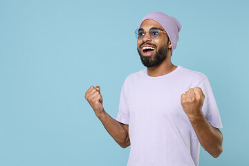 Naklejka premium Young smiling happy excited unshaven black dark-skinned african man 20s in violet t-shirt purple hat glasses do winner gesture clench fist celebrate isolated on pastel blue background studio portrait.