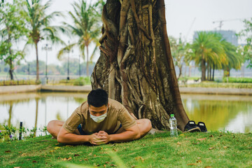 Asian man practicing meditation with wear protective mask on face, yoga exercise under big tree in...