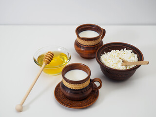 Clay jug with milk, clay cup with milk, bowl with farm cottage cheese, natural honey with honey ladle on a white table. Breakfast in earthenware. Pottery
