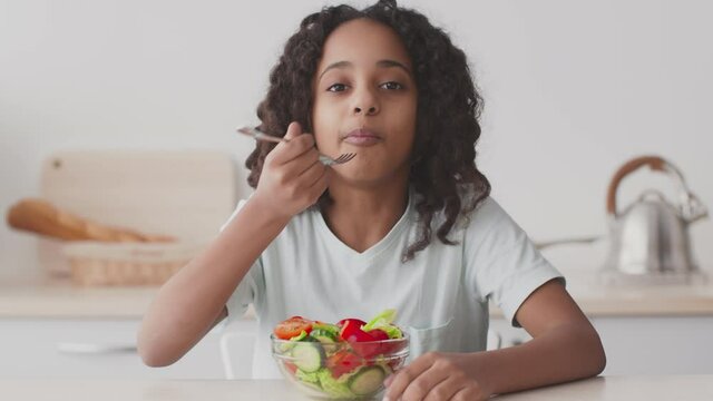 Cute african american teen girl enjoying fresh healthy vegetable salad, sitting at table at home, slow motion