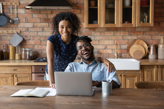 Portrait of happy loving modern day african american family couple engaged in using laptop surfing web at kitchen. Young bonding black spouses look at camera distracted of electronic paperwork on pc