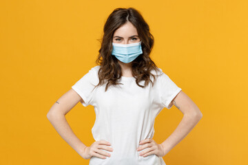 Young woman 20s in white basic t-shirt in sterile face mask to safe from coronavirus virus covid-19 during pandemic quarantine akimbo hold hands on waist isolated on yellow background studio portrait.