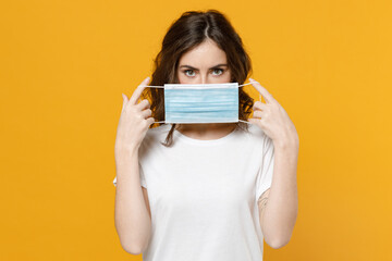 Young woman in white basic casual t-shirt hold hiding with sterile face mask from coronavirus virus covid-19 pandemic quarantine looking camera isolated on yellow orange background studio portrait.