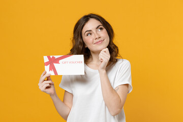 Fototapeta na wymiar Young happy dreamful pensive wistful fun caucasian woman in white basic t-shirt point index finger on gift voucher flyer mock up prop up chin look aside isolated on yellow background studio portrait.