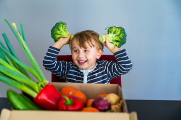 Fototapeta premium Little boy sitting at the table and taking broccoli in his hand. Set of different fresh vegetables in a cardboard box. Teaching child to healthy and varied vegitarian food.