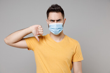 Young caucasian displeased man 20s in yellow basic t-shirt in sterile face mask to safe coronavirus covid-19 pandemic quarantine showing thumb down gesture isolated on grey background studio portrait