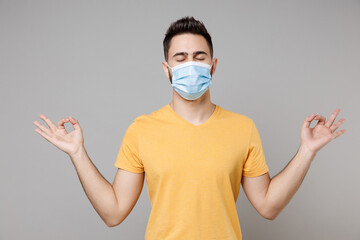 Young man in yellow t-shirt in sterile face mask to safe coronavirus covid-19, pandemic quarantine hold hands in yoga gesture meditating, try to calm down isolated on grey background studio portrait.