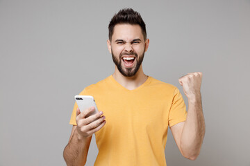 Young caucasian smiling happy bearded attractive man in casual yellow t-shirt hold mobile cell phone chat in social network do winner gesture clench fist isolated on grey background studio portrait.