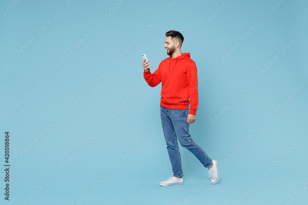 Wall mural Full length side profile view of young caucasian smiling bearded attractive man 20s wear casual red orange hoodie holding mobile cell phone walking going isolated on blue background studio portrait - Wall murals