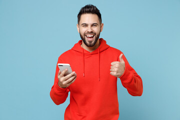 Young caucasian smiling happy bearded man 20s in casual red orange hoodie hold mobile cell phone texting show thumb up gesture isolated on blue background studio portrait People lifestyle concept