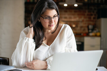 Indoor shot of attractive plus size young black haired woman in eyeglasses and white blouse sitting in front of open laptop watching video course online using wireless earplugs, having interested look