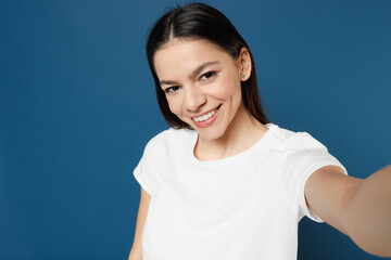 Young smiling attractive beautiful cheerful cute happy latin woman 20s in white casual basic t-shirt doing selfie shot on mobile phone looking camera isolated on dark blue background studio portrait