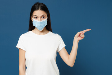 Young latin woman 20s in white t-shirt sterile face mask to safe coronavirus covid-19 pandemic quarantine point index finger aside on workspace area isolated on dark blue background studio portrait.