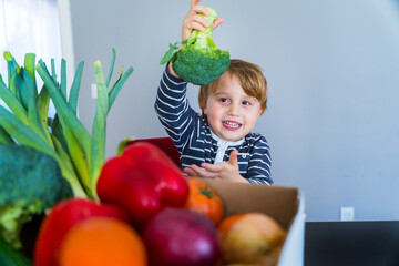 Fototapeta na wymiar Little boy sitting at the table and taking broccoli in his hand. Set of different fresh vegetables in a cardboard box. Teaching child to healthy and varied vegitaria food.