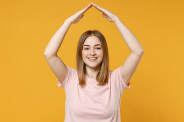 Young smiling friendly happy woman 20s, nude make up wearing casual basic pastel pink t-shirt holding folded hands above head like roof, stay home isolated on yellow color background studio portrait