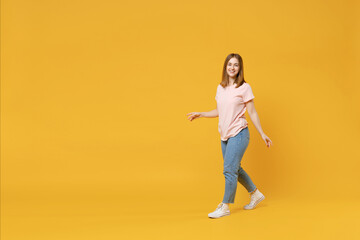 Full length of young friendly smiling student woman 20s with nude make up wearing casual basic pastel pink t-shirt, jeans looking camera, walking going isolated on yellow background studio portrait.