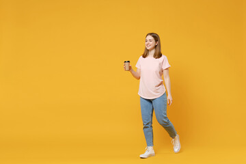 Full length of young caucasian student woman 20s wearing casual basic pastel pink t-shirt, jeans holding paper cup of tea drinking coffee walk going isolated on yellow background studio portrait.