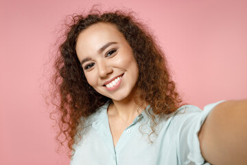 Close up young black african fun american smiling happy curly woman 20s in blue shirt doing selfie shot on mobile phone talking by video call isolated on pastel pink color background studio portrait