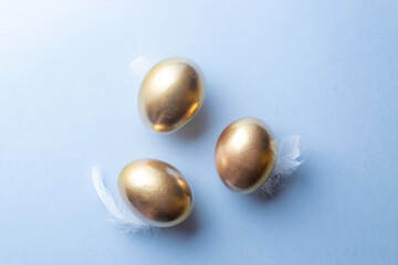 Golden easter colour eggs in basket with white feathers on pastel blue background in Happy Easter decoration. Spring holiday top view concept.