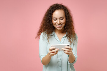Young black african curly friendly smiling fun happy woman 20s wearing casual blue shirt hold in hand using mobile cell phone playing games isolated on pastel pink color background studio portrait.