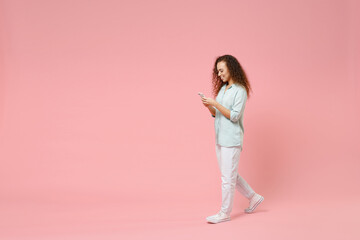 Full length young black african fun happy smiling curly student woman 20s in blue shirt holding mobile cell phone chatting browsing internet walk isolated on pastel pink background studio portrait.
