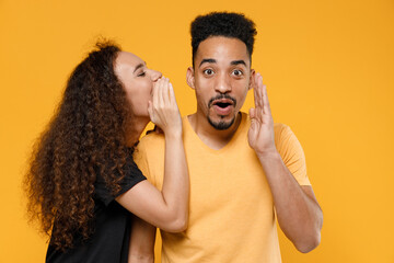 Young couple two friends family african shocked man woman in black t-shirt whisper gossip and tell secret behind hand share news with guy spreading hand isolated on yellow background studio portrait.