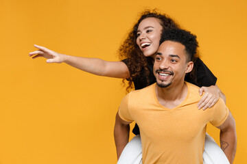 Young couple two friends together family smiling happy cheerful african man woman in yellow black tshirt giving piggyback ride to joyful, sitting on back isolated on orange background studio portrait.