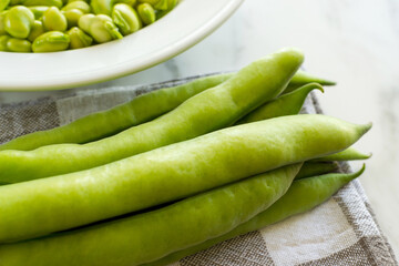 Detail of bean pods on tablecloth and plate full of beans in the background, on marble table in the kitchen.