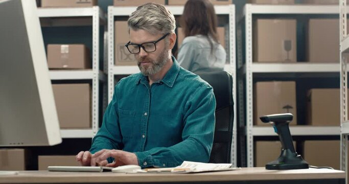 Warehouse director with gray hair and beard with glasses works at computer sitting on background of cardboard boxes.