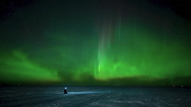 Person sitting on a frozen snow covered lake watching active green Aurora.  The subject is wearing a white head lamp.

