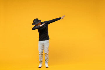 Full length young fun african man 20s wear stylish black hat shirt eyeglasses doing dab hip hop dance hands move gesture youth sign hiding covering face isolated on yellow background studio portrait