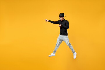 Fototapeta na wymiar Full length side view of young active fashionable african man in stylish black hat shirt eyeglasses jump high point index finger aside on workspace area isolated on yellow background studio portrait.