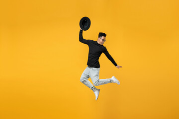 Plakat Full length of young active fashionable overjoyed fun happy african man in stylish black shirt eyeglasses jump high with outstretched hands took off hat isolated on yellow background studio portrait.