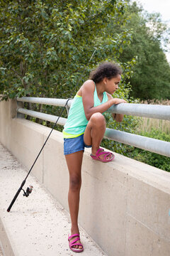 Girl leaning against the metal rail of a country bridge along with her fishing rod