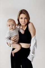 Happy young mother with lovely baby girl posing in the studio. Stylish, beautiful woman hold little daughter in arms, looking confidently at the camera, smiling