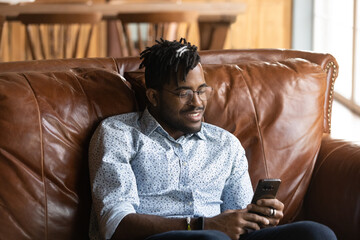 Fototapeta premium Passionate gamer. Joyful millennial african guy student relax at home sit on couch play smartphone based online game. Young black man hipster addicted to internet spend free time using mobile phone