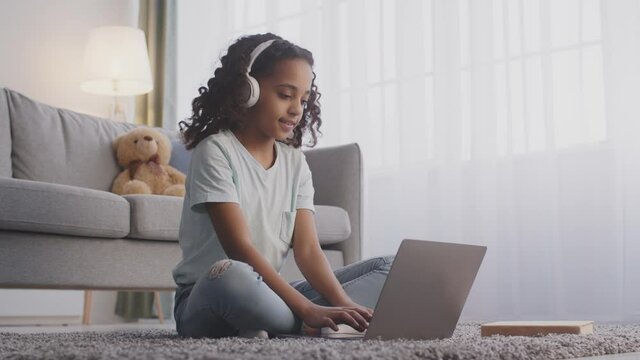 Teen black girl in headphones typing on laptop, communicating with teacher via video chat, sitting on floor at home
