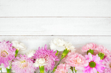 Bottom border of pink and purple flowers with mums, daisies and carnations against a white wood background. Copy space.